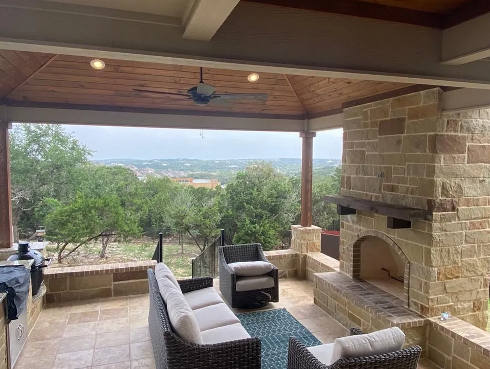 A patio with a fireplace , couch , chairs and a ceiling fan built by Fine Patio Design San Antonio TX