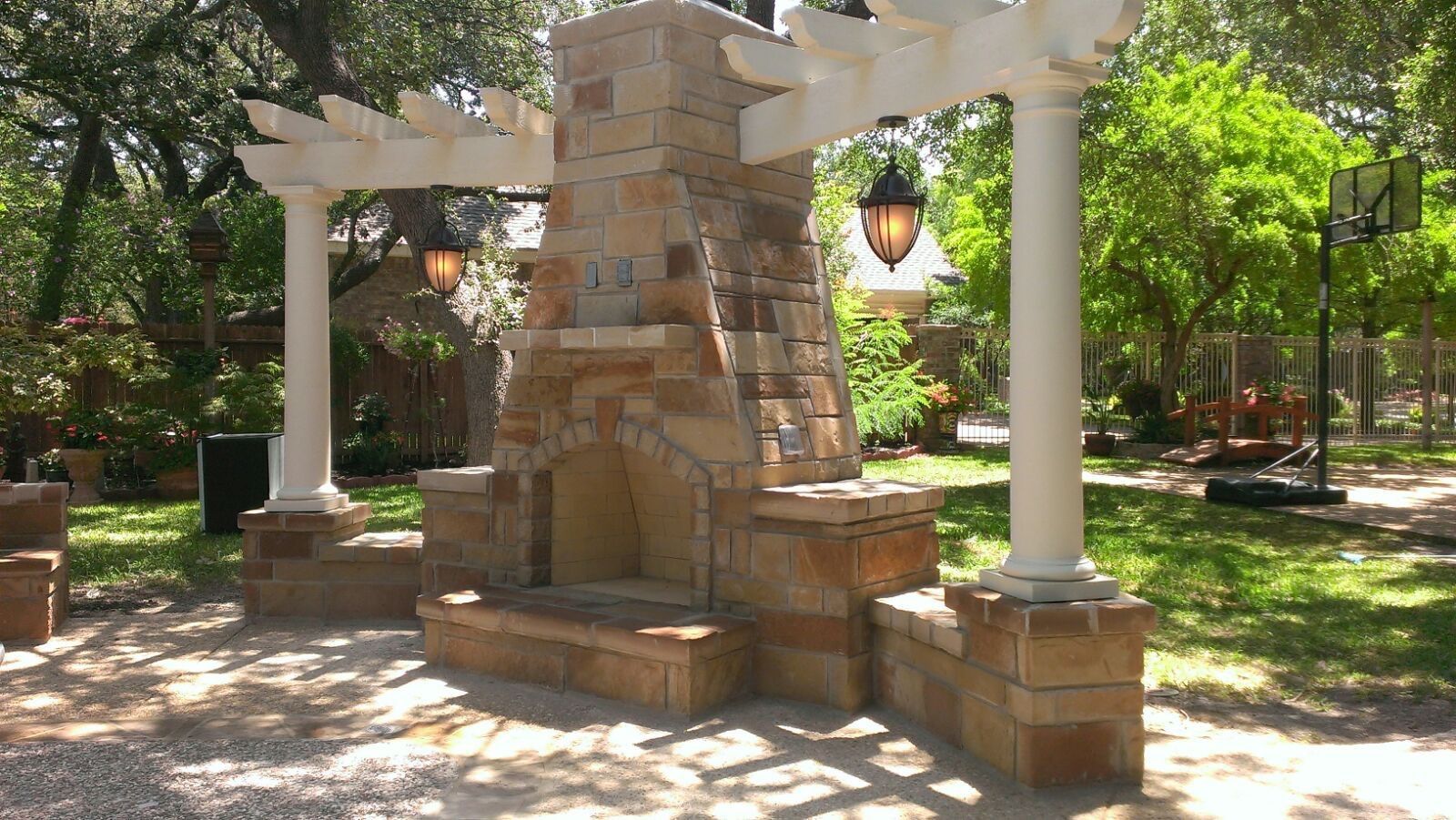 A stone fireplace under a white pergola in a backyard built by Fine Patio Design San Antonio TX