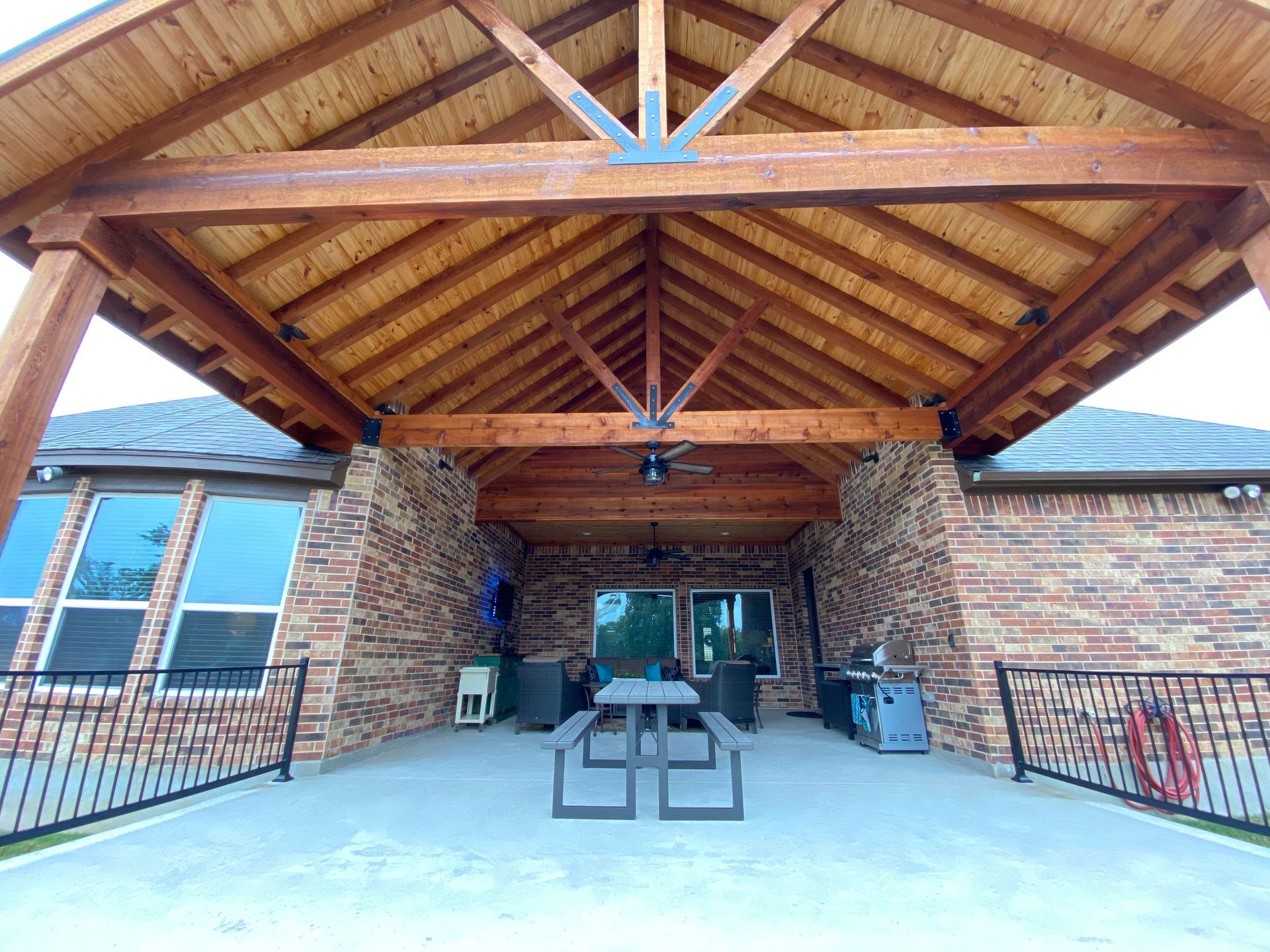 A picnic table under a wooden roof in front of a brick building built by Fine Patio Design San Antonio TX
