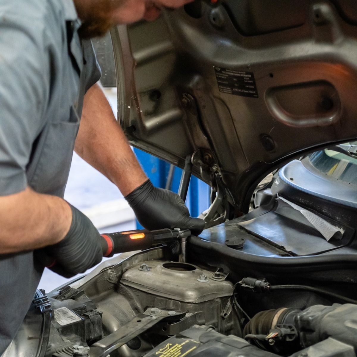 Mechanic working on a car engine with a wrench. Wearing gloves, gray shirt, and in a garage setting | Fort Meade Auto Center