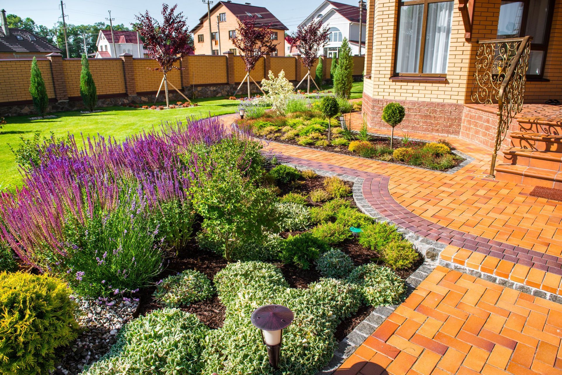 A sunlit garden path made of orange bricks curves past purple flowers and small green shrubs beside a brick house.