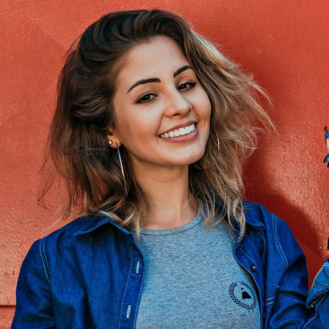 A smiling person with shoulder-length wavy hair wears a grey t-shirt and blue denim shirt against a vibrant orange wall.