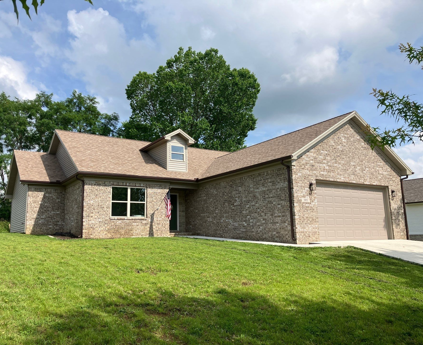 Single-story brick house with a beige garage, front lawn, and trees under a partly cloudy sky
