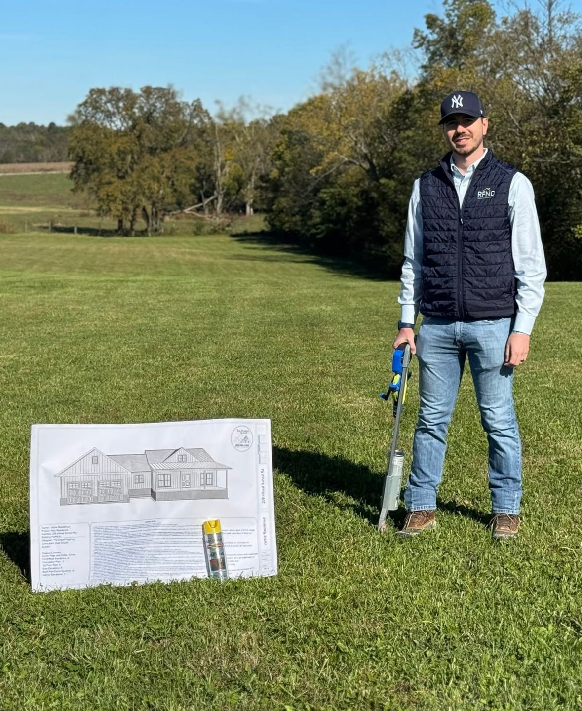 Man standing on a grassy field beside a house plan board, holding golf clubs