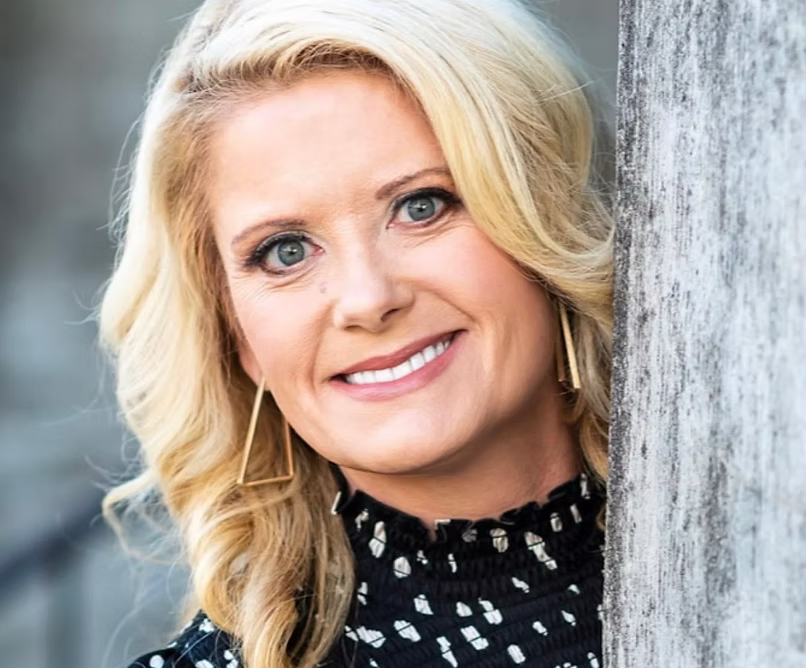 Smiling blonde woman leaning against a gray wall, wearing a black patterned top