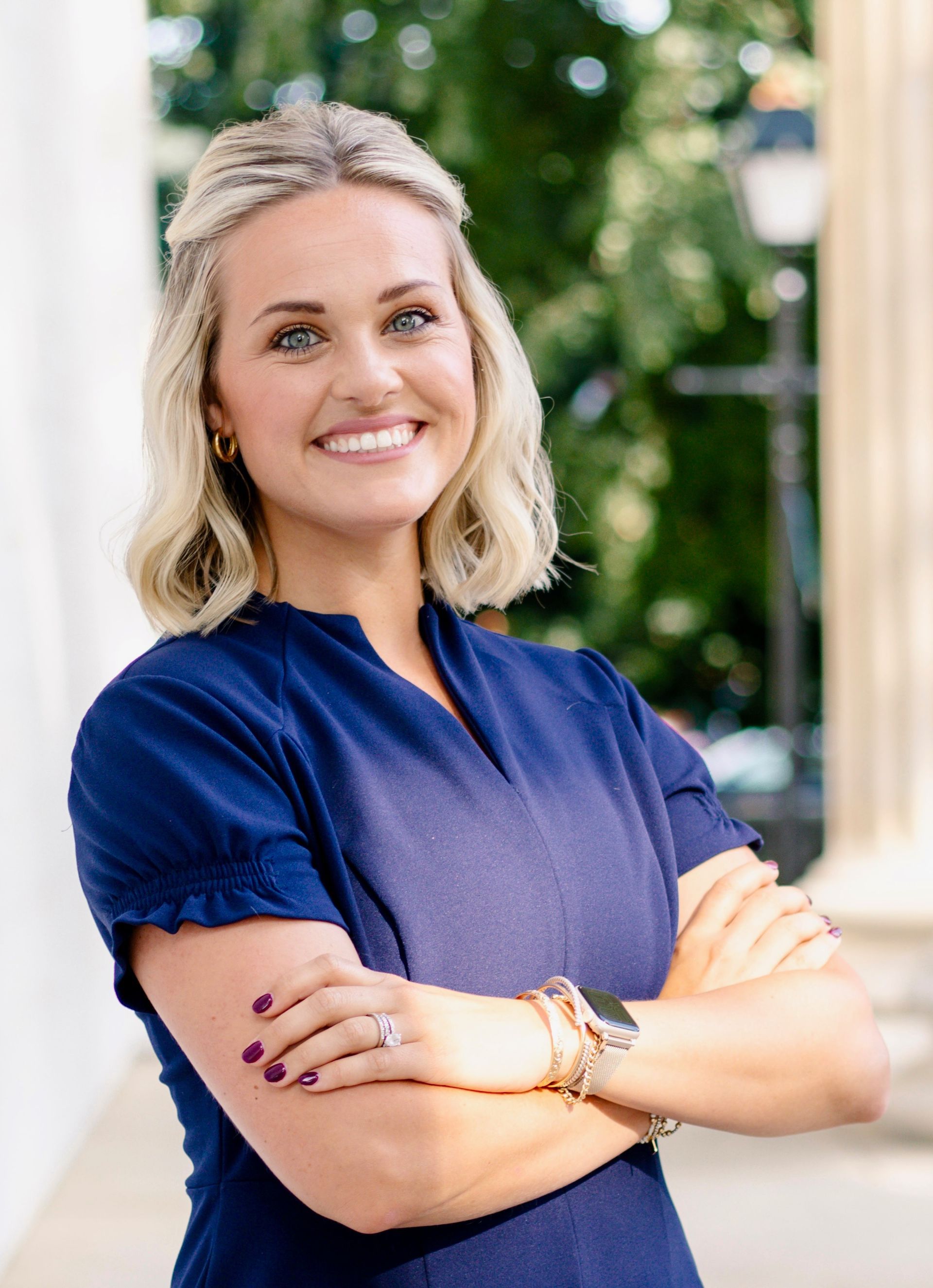 Smiling woman with blonde hair in a navy blouse, standing outdoors with arms crossed.