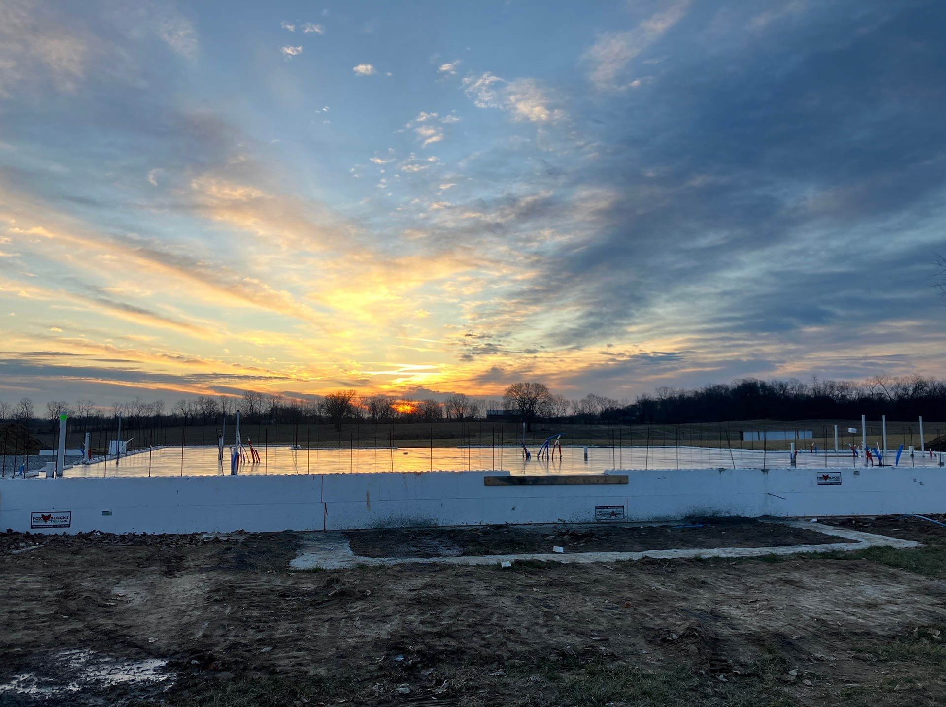 Sunset over a snowy construction site with reflective water and a dramatic cloud-filled sky