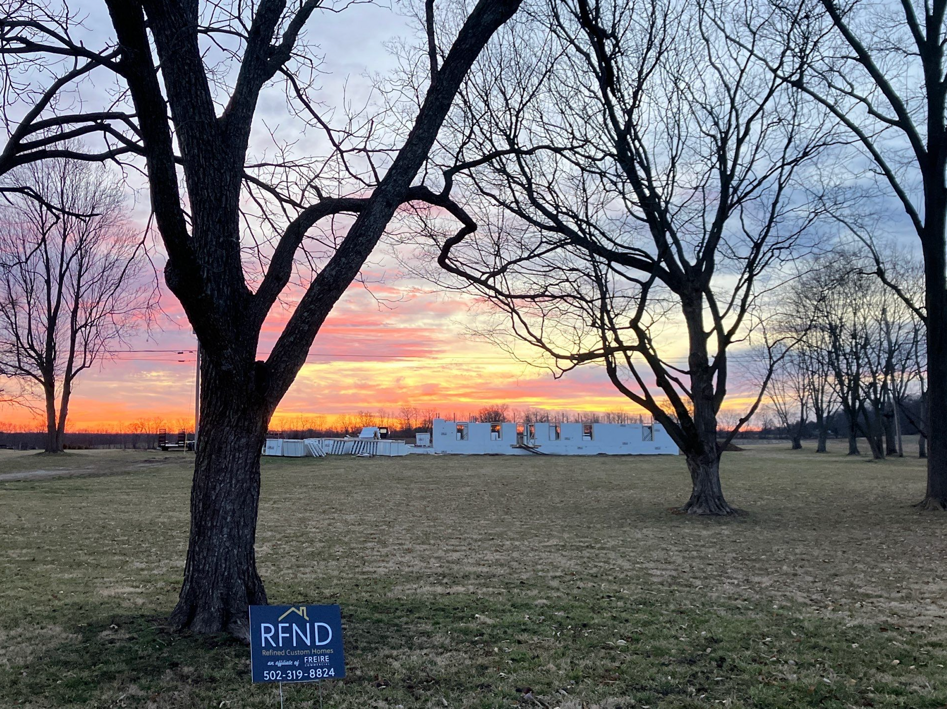 Bare trees silhouetted against a pink-orange sunset over a grassy field with a small RFND sign in front