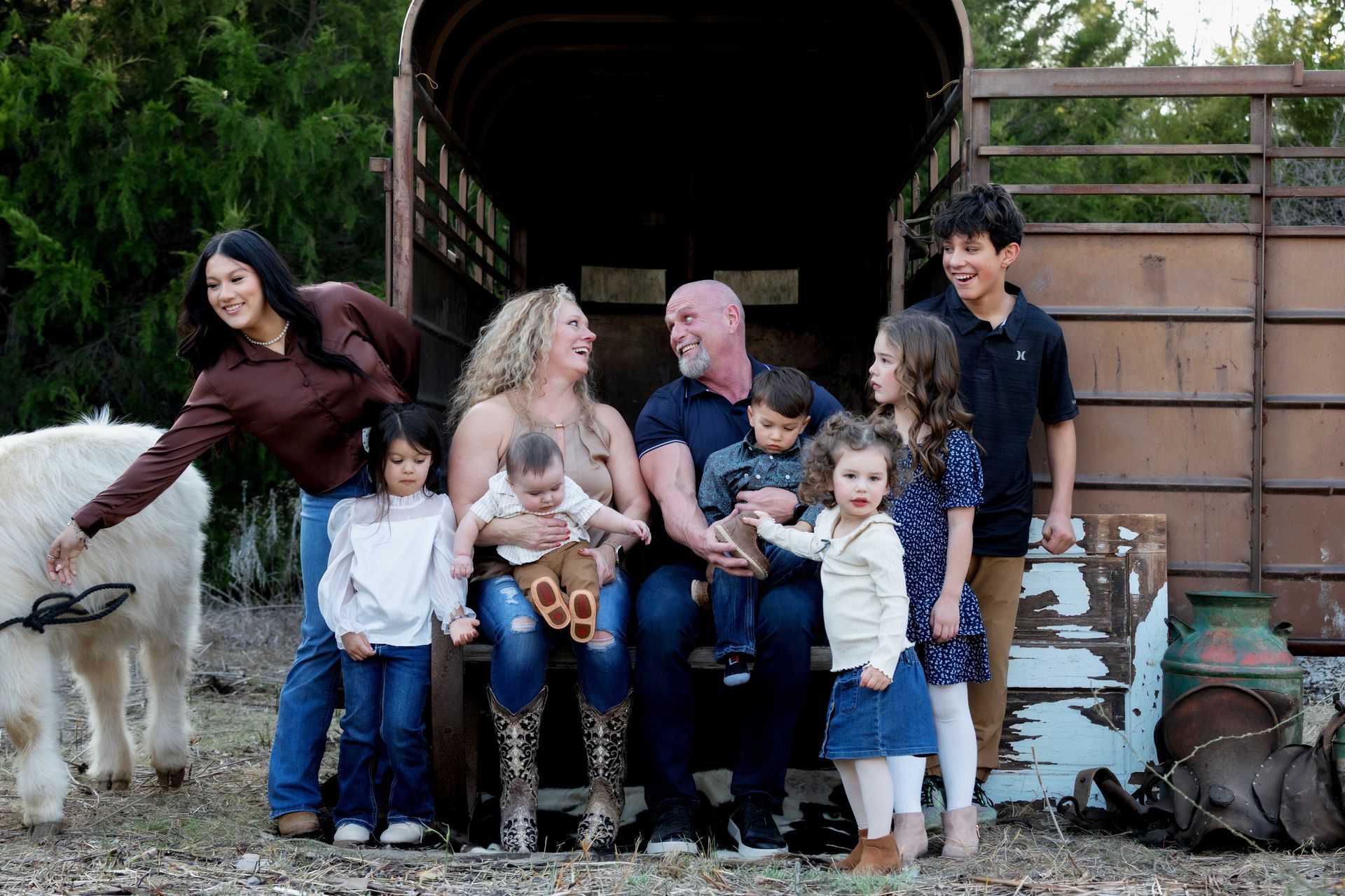 Family sitting with two goats in front of a barn, smiling children and adults gathered outdoors