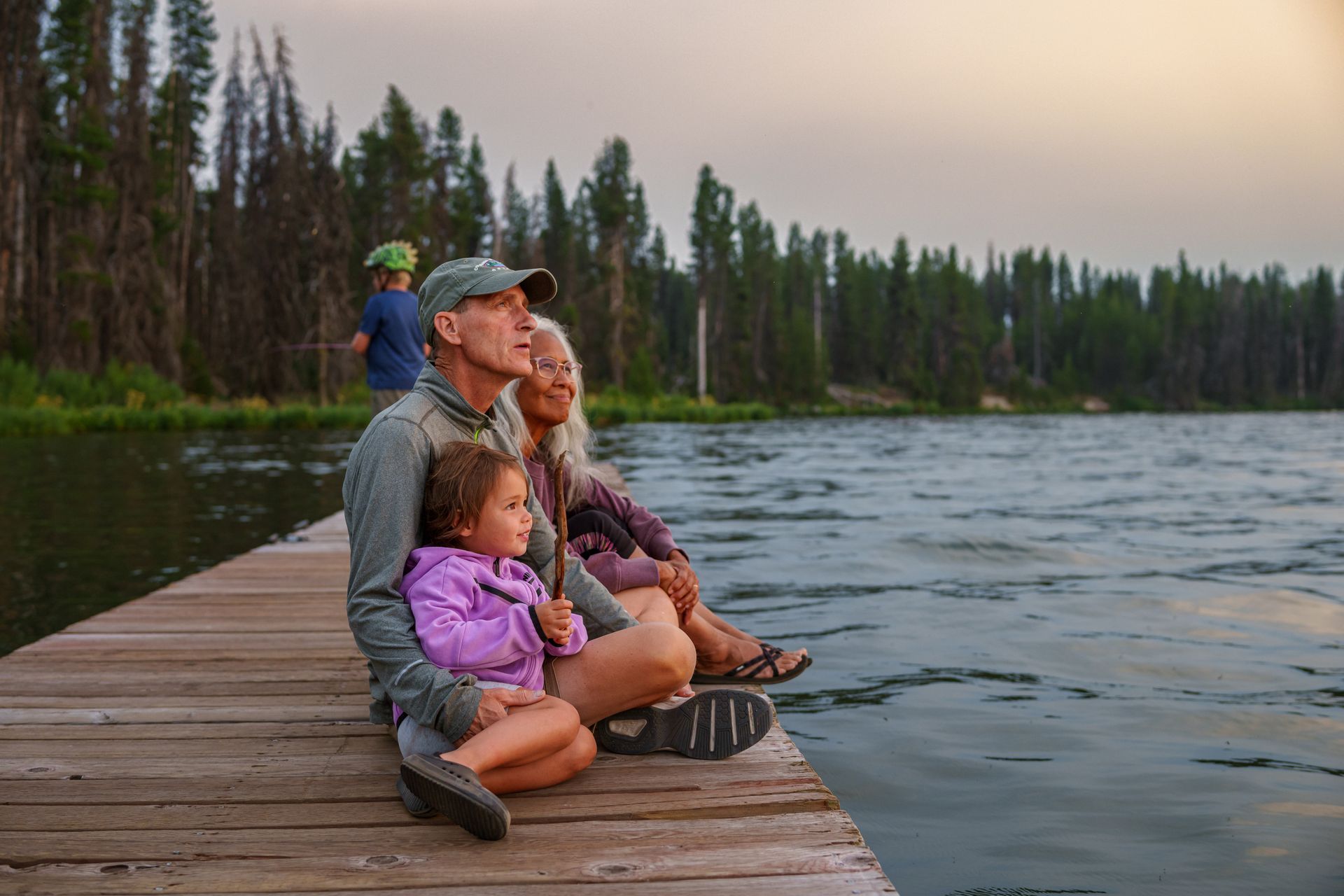 Family sitting on a wooden dock by a lake at sunset, looking out over the water and trees.