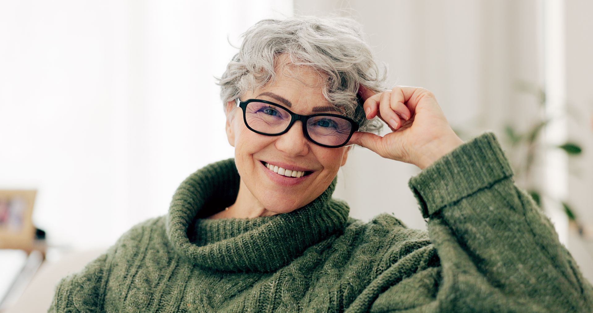 Smiling person in a green sweater adjusting glasses indoors by a bright window
