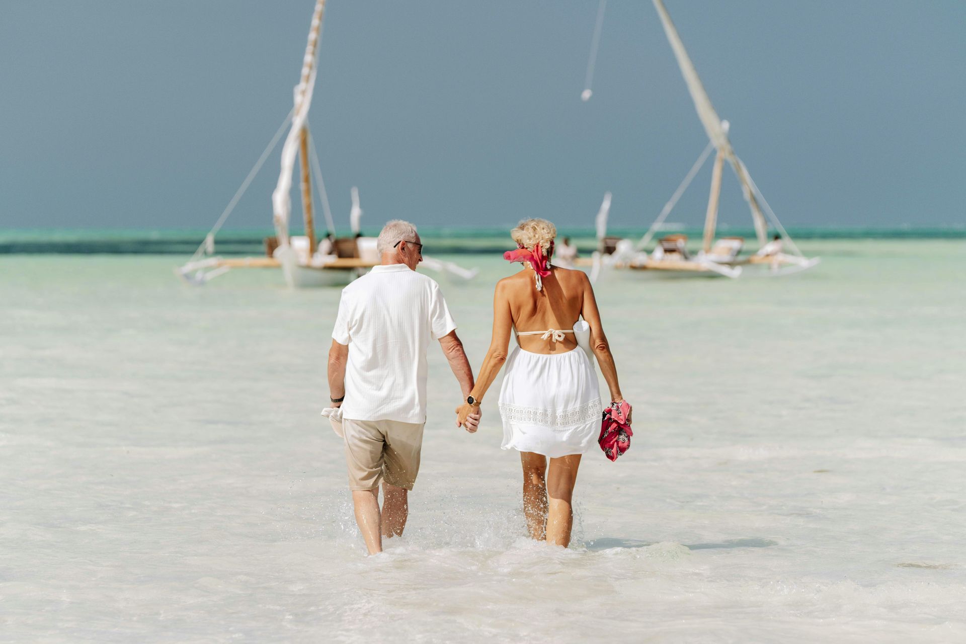 Couple walking hand in hand through shallow turquoise water near sailboats on a tropical beach
