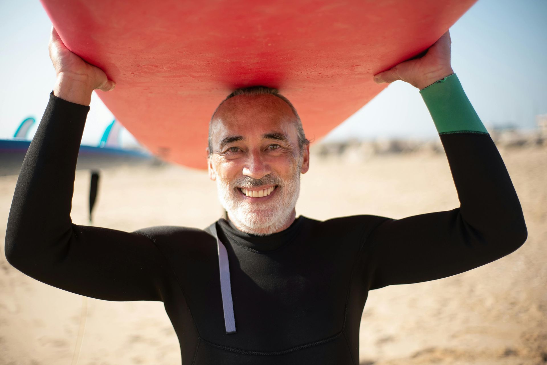 Man in black wetsuit holding a red surfboard overhead on a sandy beach, smiling at the camera