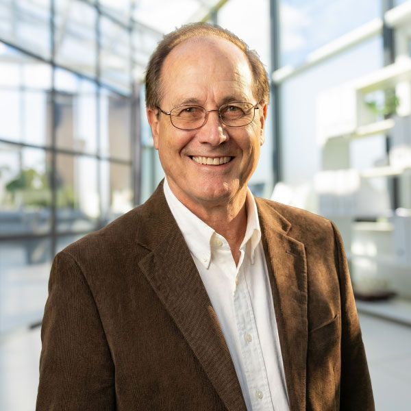 A person wearing a brown corduroy blazer and glasses smiles in a bright, modern office space with large windows.