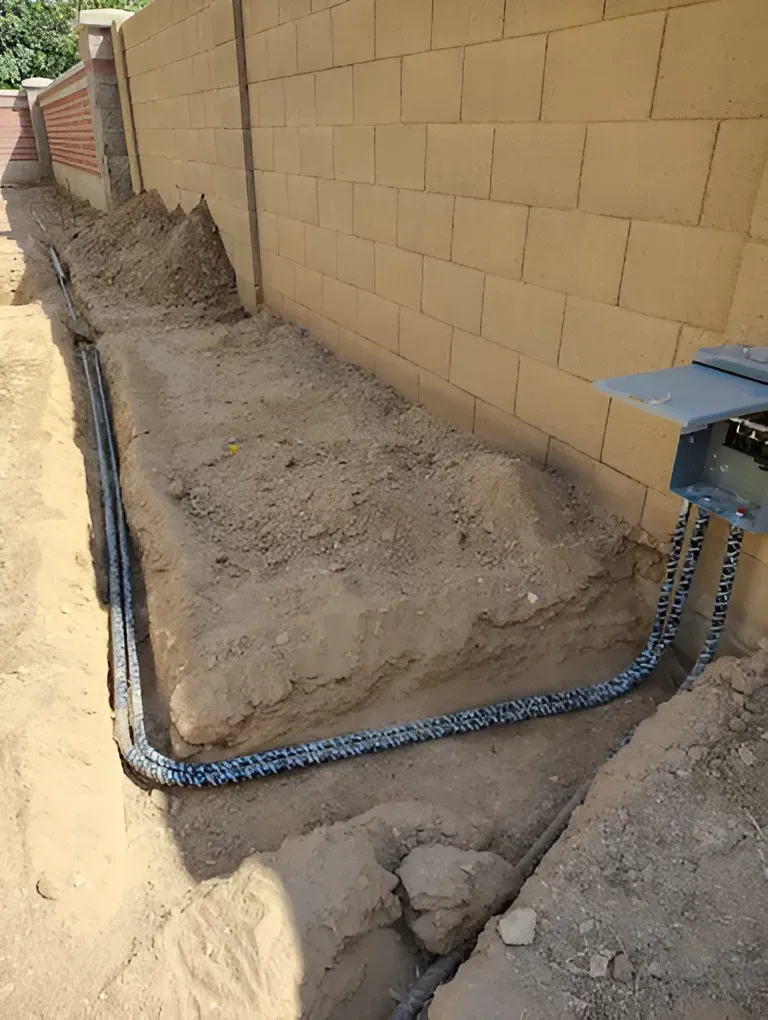 Trench with black conduit running alongside a tan brick wall and electrical box.