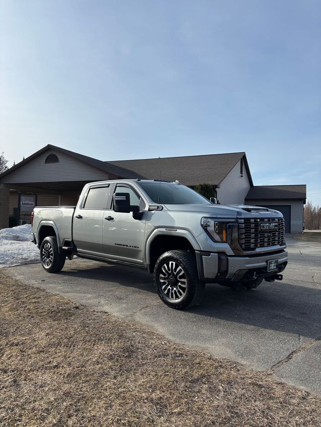 Silver GMC truck parked in front of a house on a sunny day. The truck has black wheels and a front-mounted accessory.