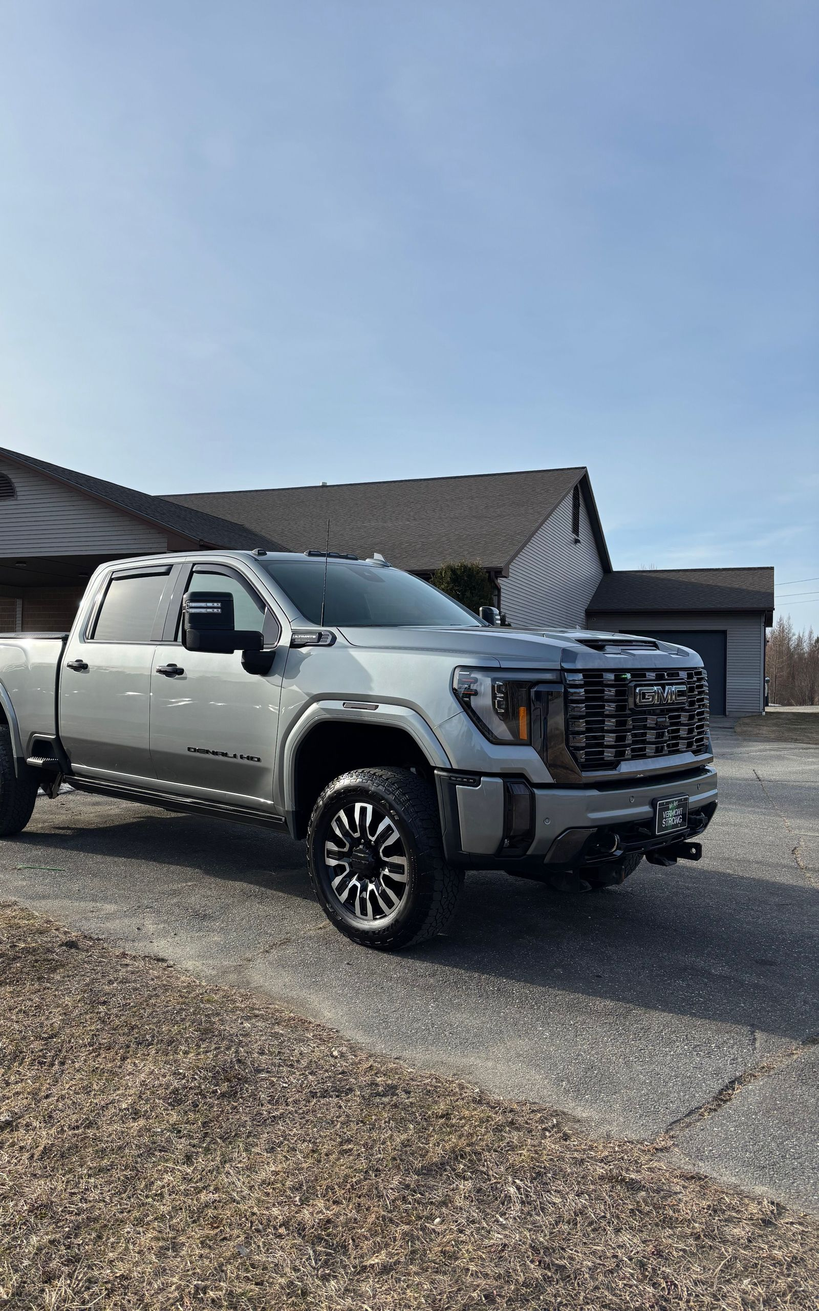 Silver GMC truck parked in front of a house on a sunny day. The truck has black wheels and a front-mounted accessory.