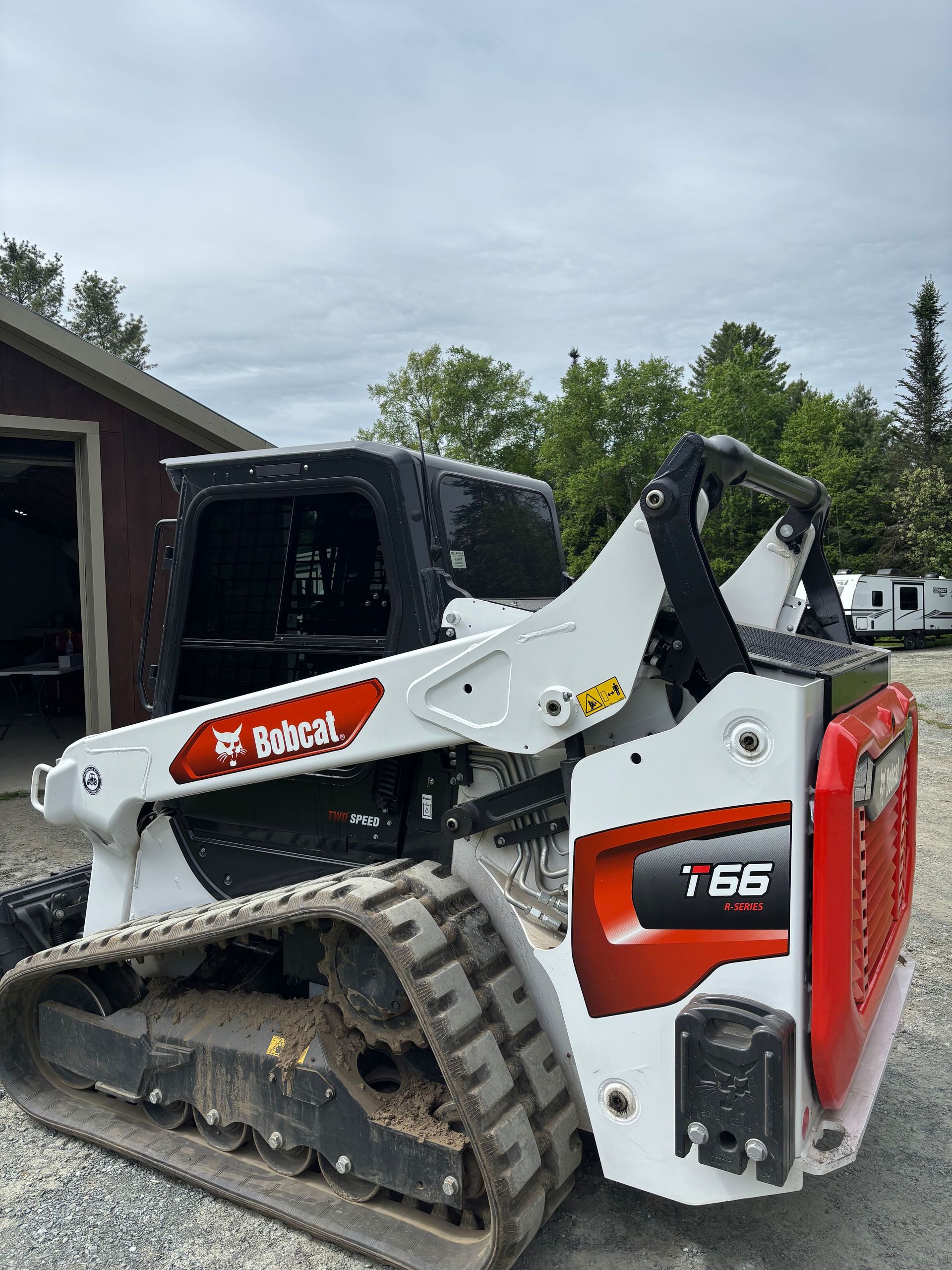 A white and red Bobcat T65 compact track loader sits outside, next to a building and trees, under a cloudy sky.