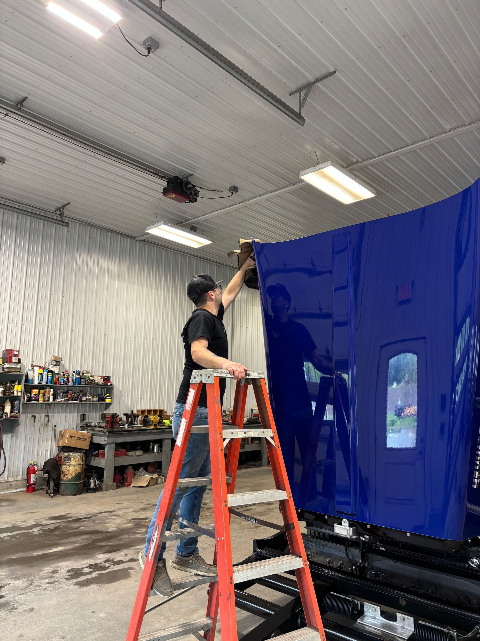 Person on a ladder applying a blue vinyl wrap to a vehicle hood inside a garage.