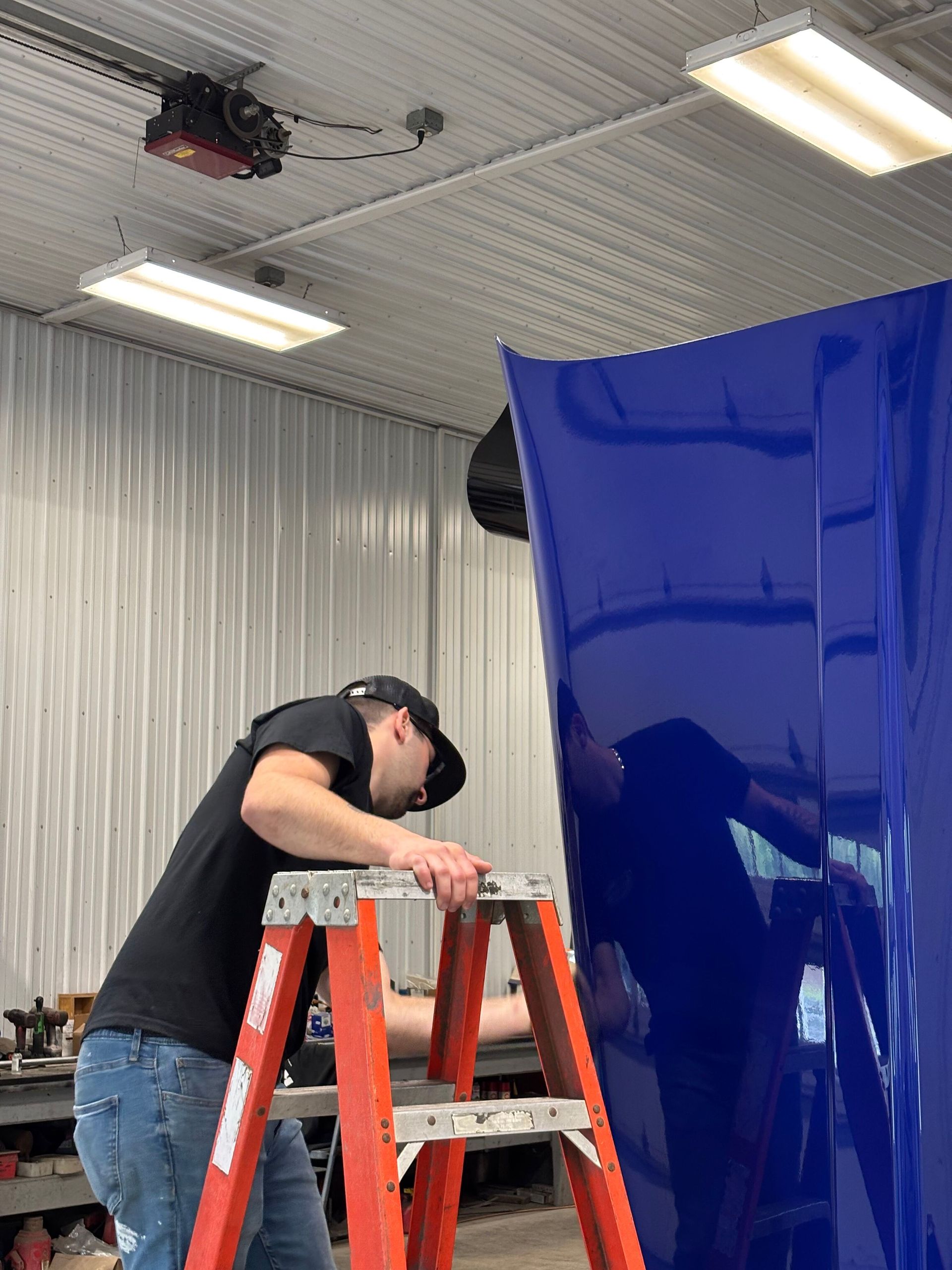 Man on a ladder inspects a large, blue, car hood inside a garage, with the hood taking up most of the frame.