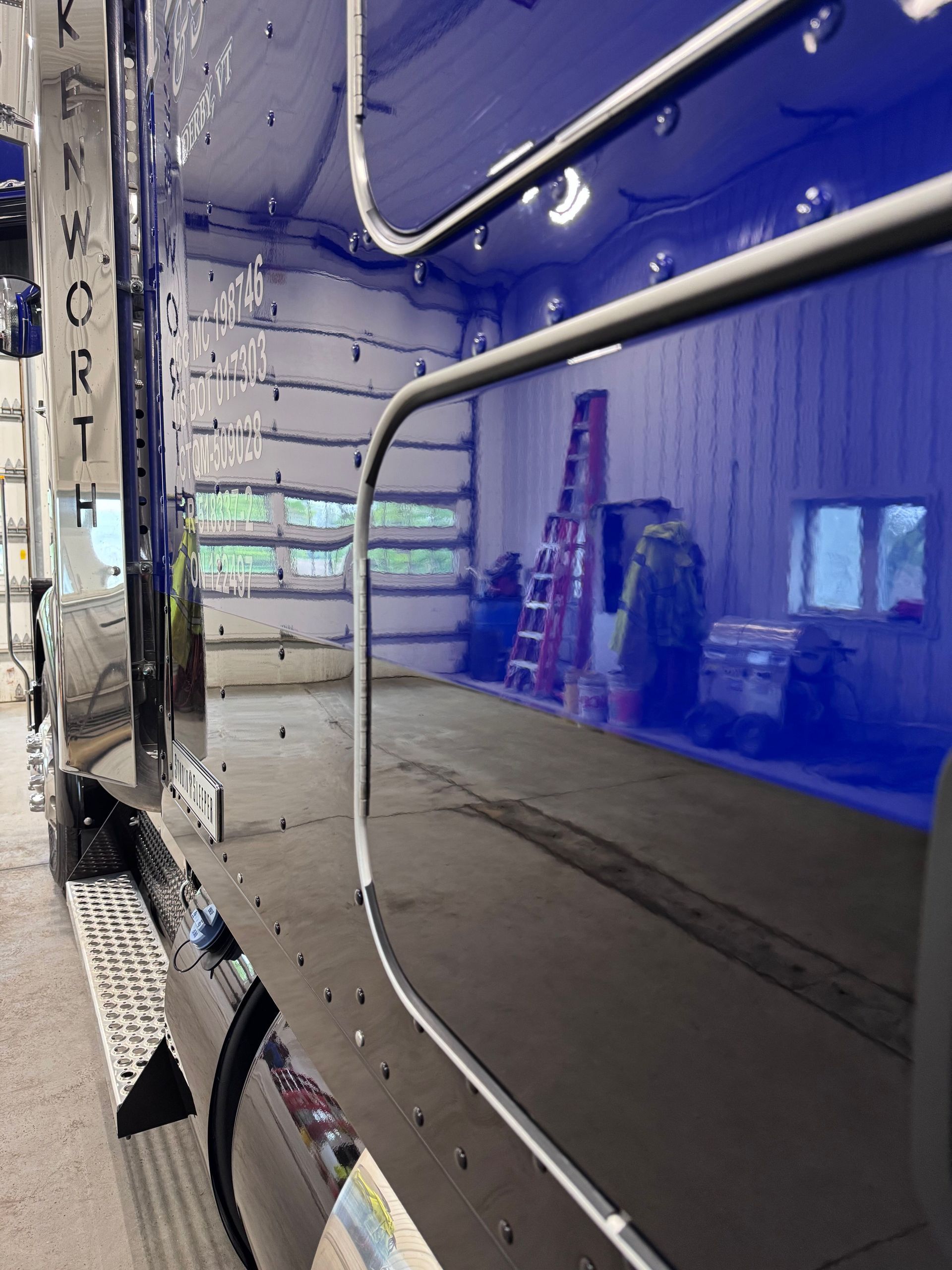 Shiny black Kenworth truck side reflecting a garage interior with a red ladder, window, and other objects.