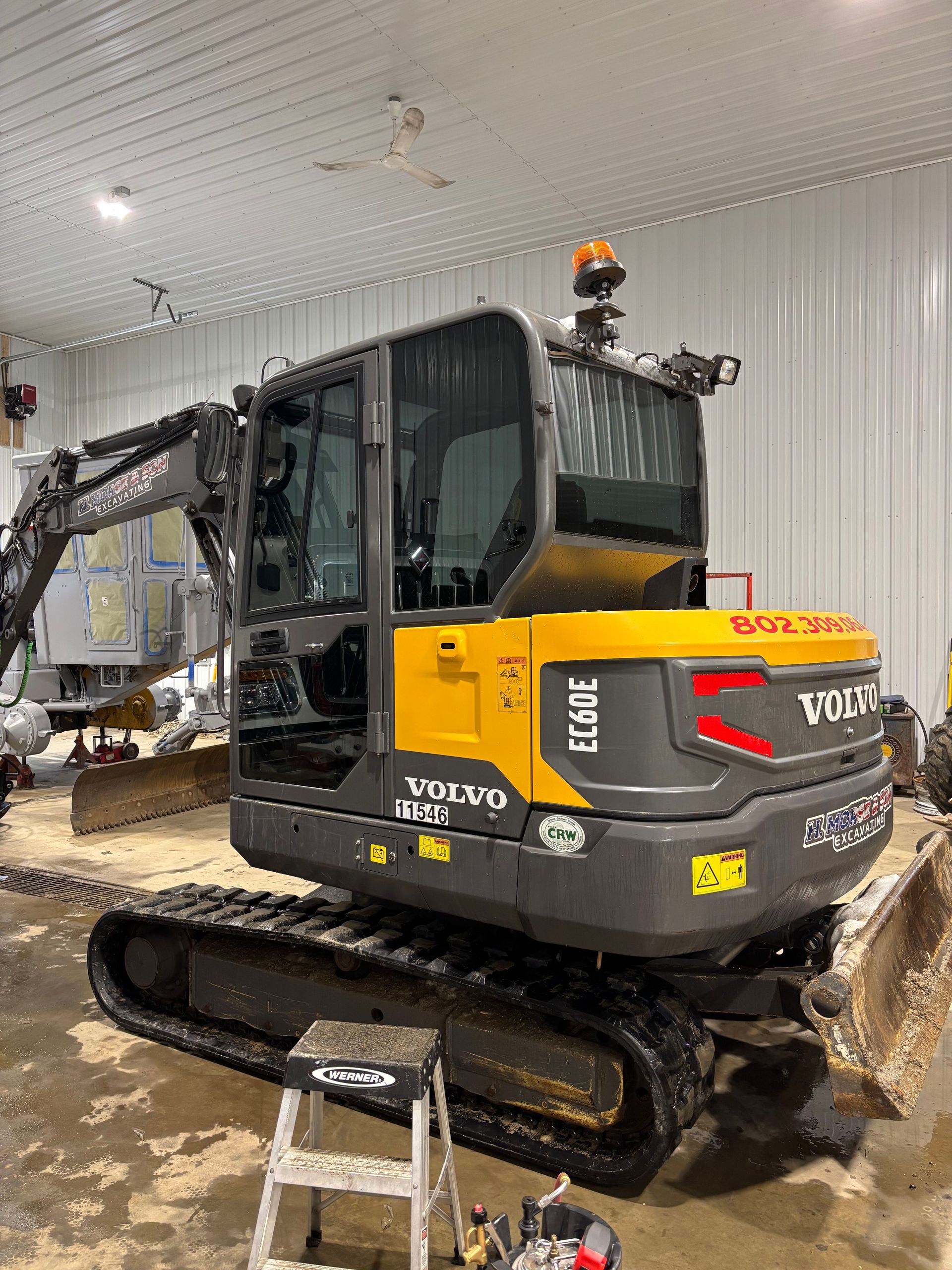 Yellow and black Volvo EC60E excavator inside a garage. The machine has a cab, tracks, and a raised arm.