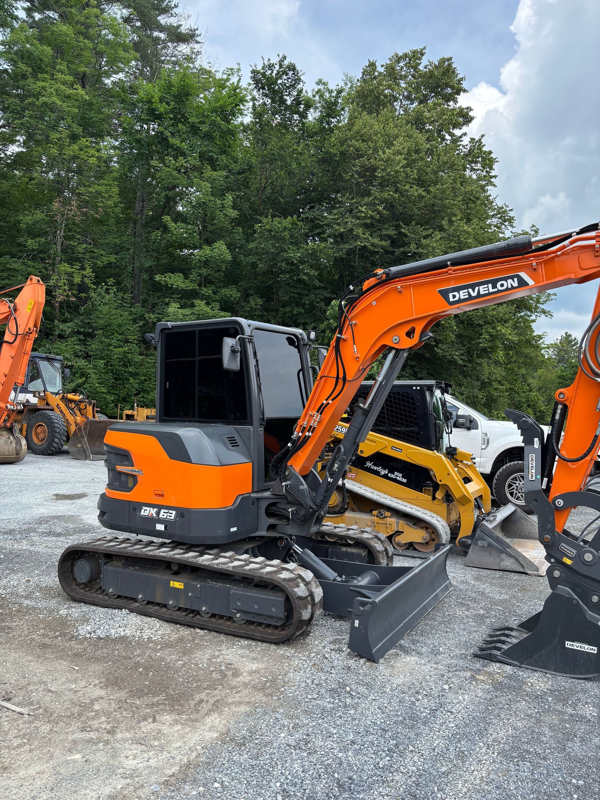 Orange and black excavator parked outdoors, with a pile of wood and trees in the background.