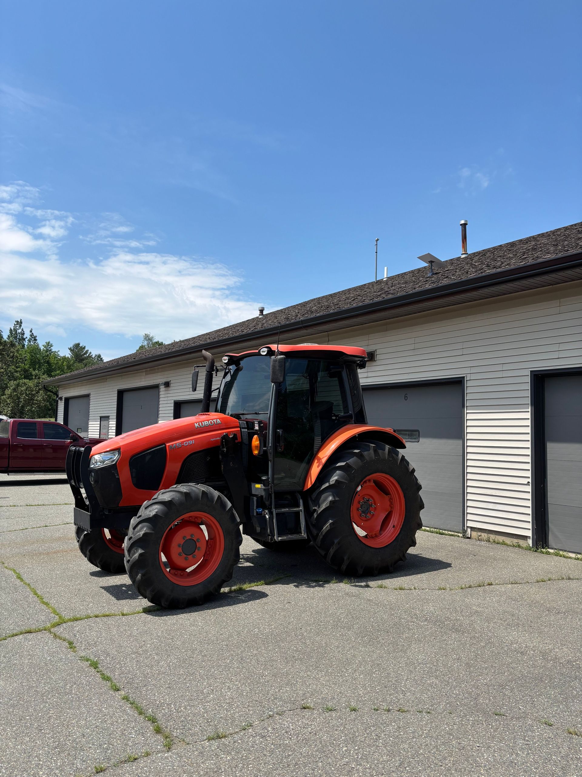Red and black Kubota tractor parked in front of a light-colored building with closed garage doors on a sunny day.