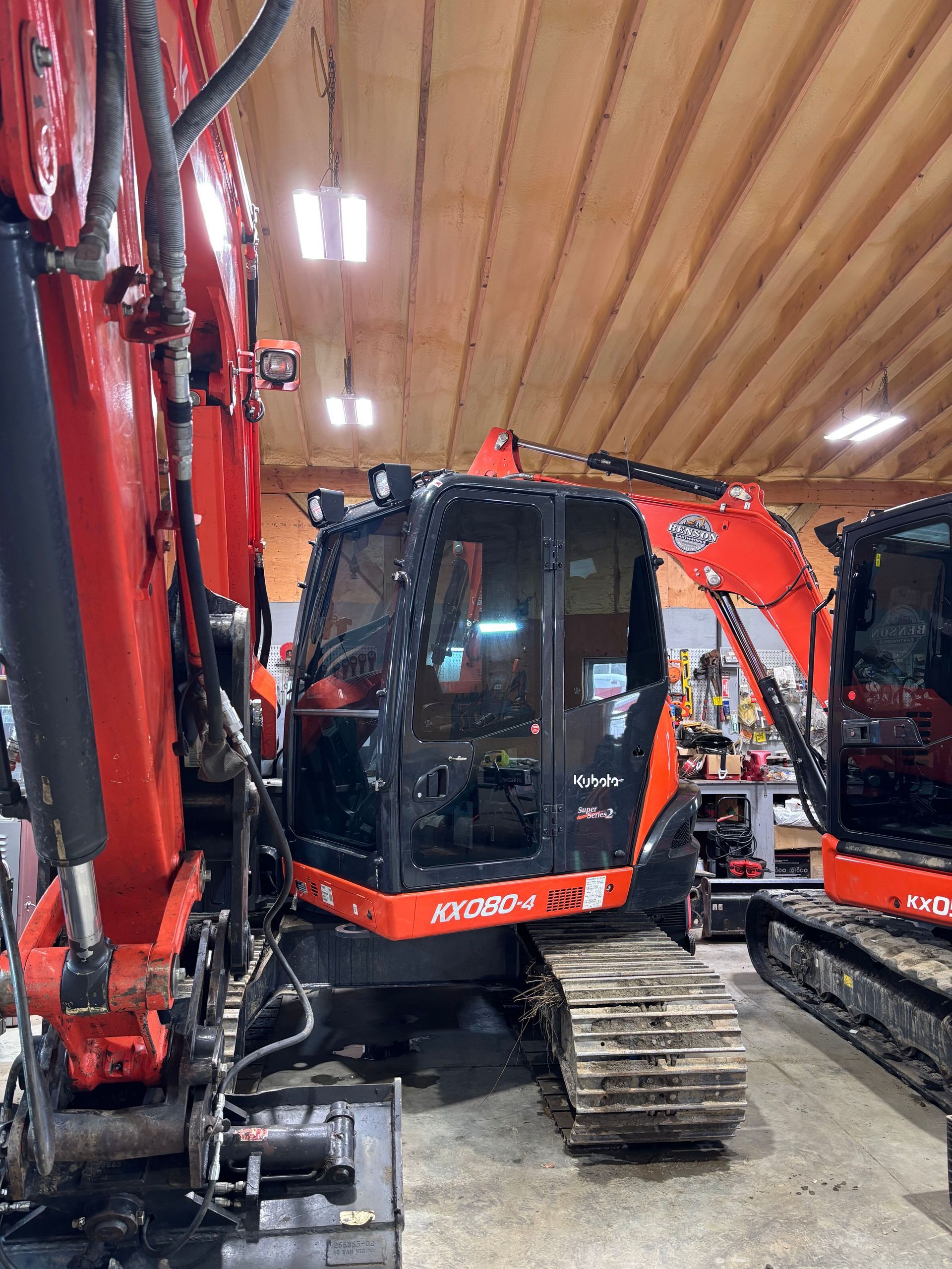 Red and black Kubota excavator inside a warehouse. The cab has black windows. Another excavator is partly visible on the right.