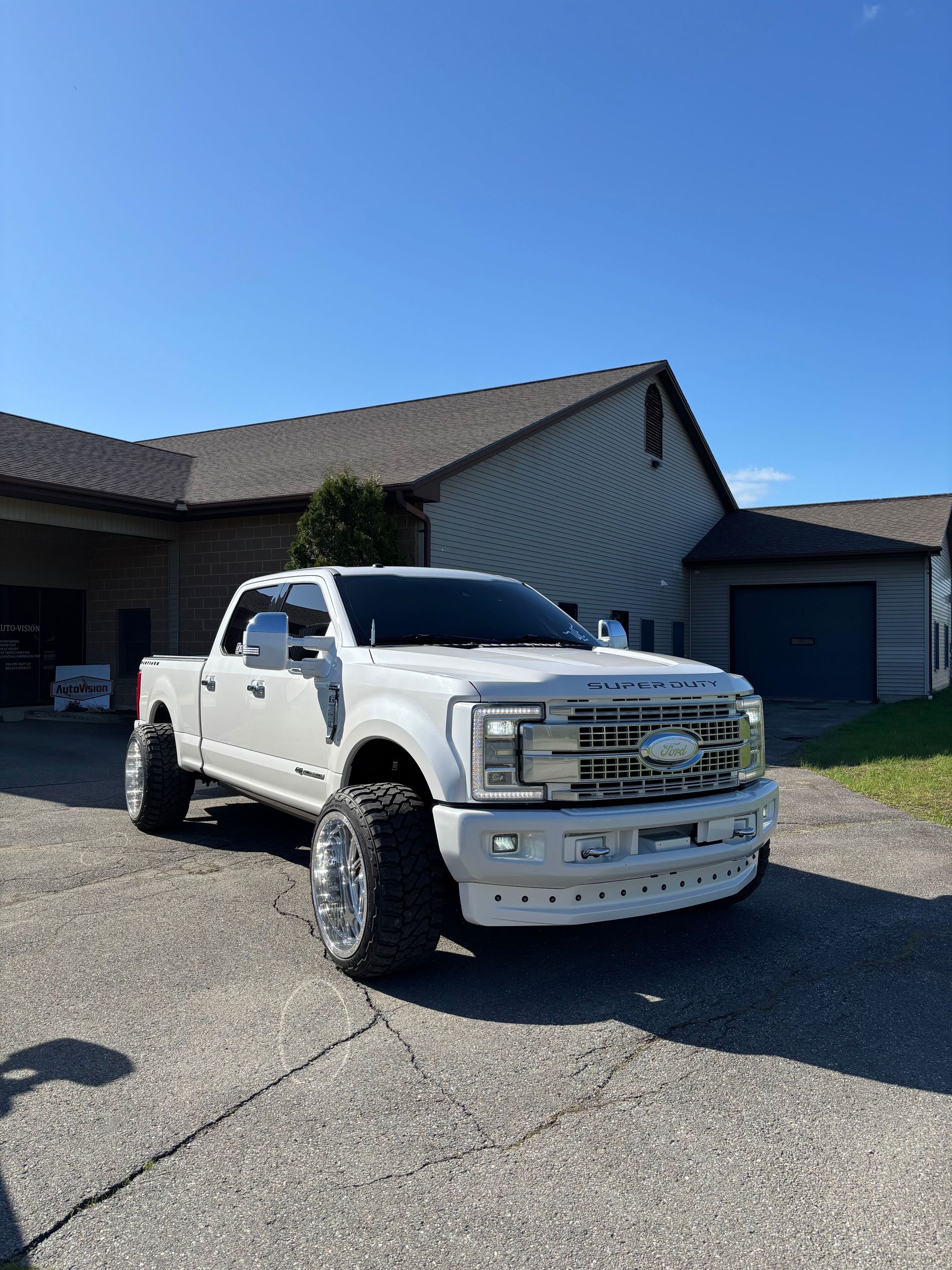 White Ford truck parked on a paved area in front of a building on a sunny day. The truck has large chrome wheels.