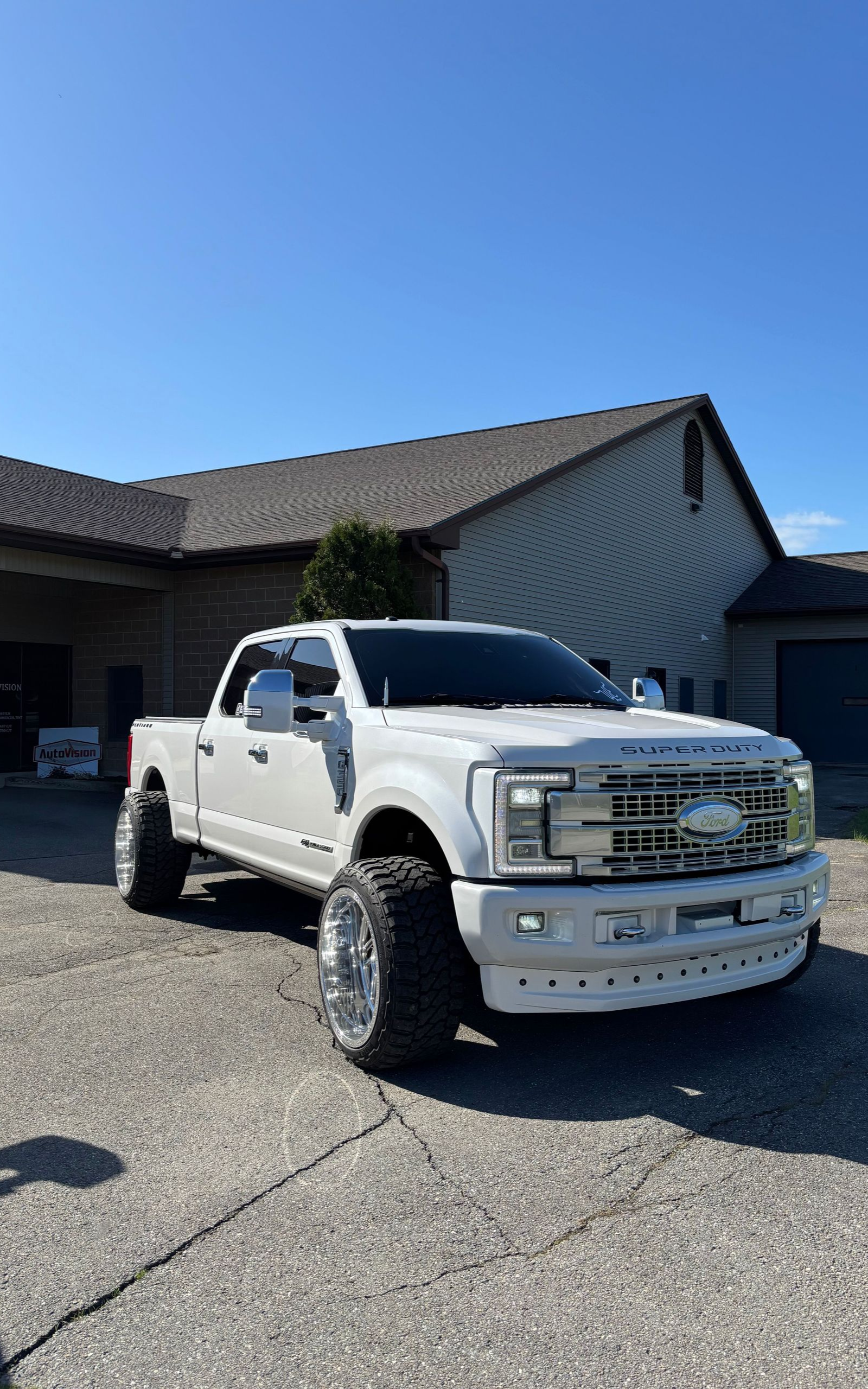 White Ford truck parked on a paved area in front of a building on a sunny day. The truck has large chrome wheels.