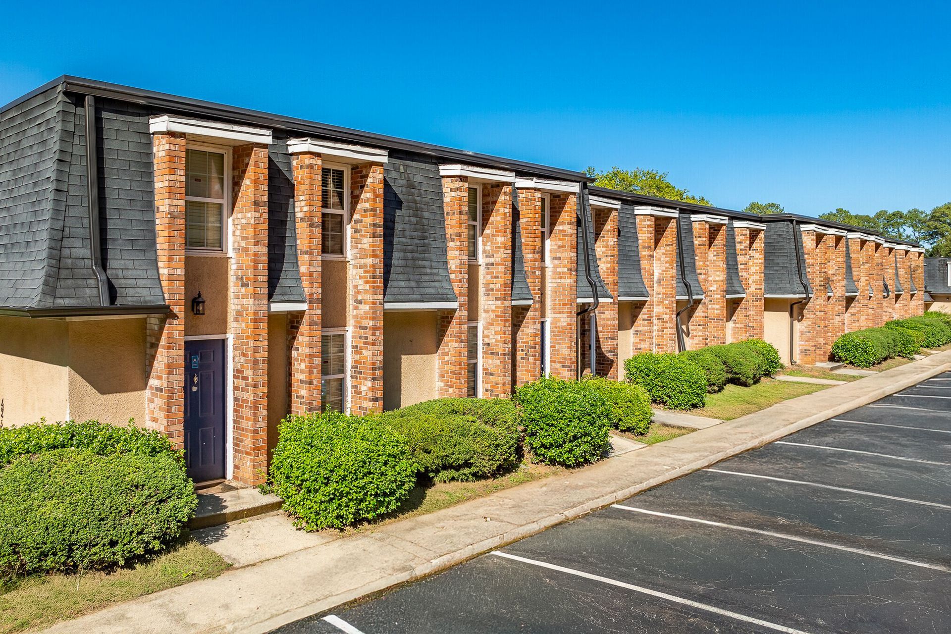Row of brick townhouses with black roofs, shrubs, and parked cars on a sunny day.
