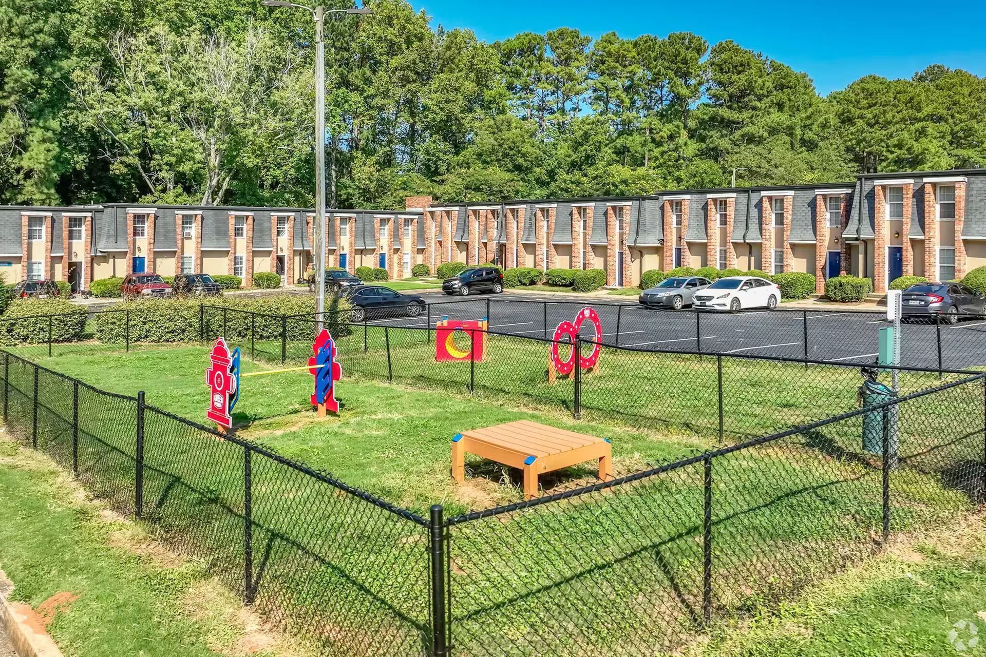 A fenced-in playground with lawn, play structures, and a row of apartments in the background.