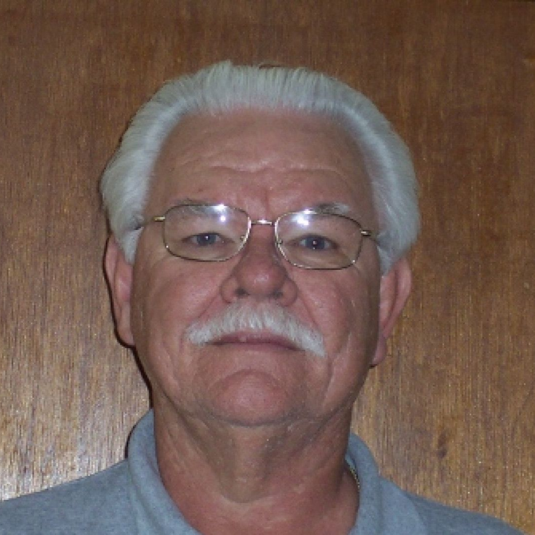 Man with glasses and white hair and mustache in front of a wooden wall.