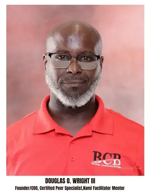 A man with a graying beard and glasses, wearing a red collared shirt with a BCB logo, on a soft-focus reddish background.