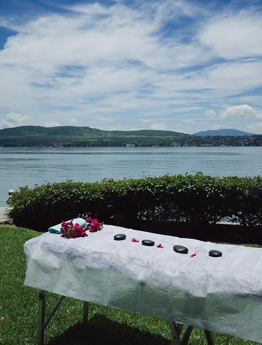 Mesa de masaje con piedras y flores con vistas a un lago y montañas en un día soleado.