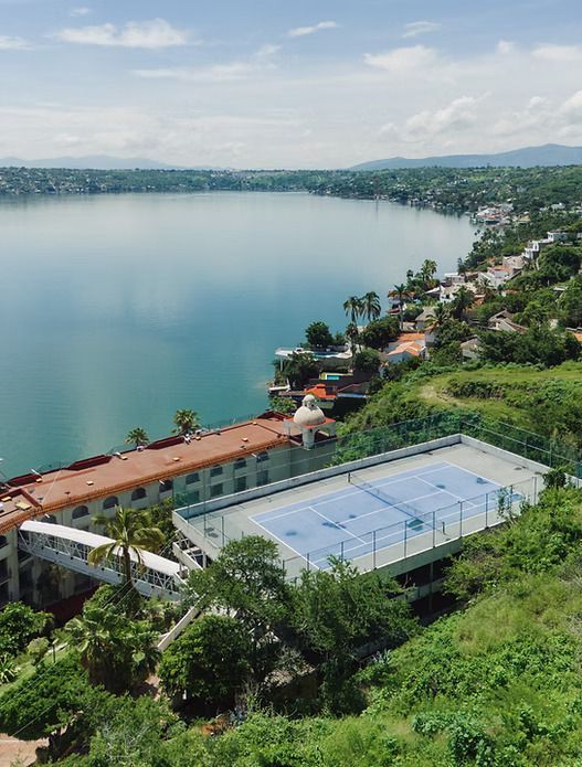 Cancha de tenis con vista a un lago y a un pueblo en la ladera bajo un cielo soleado.