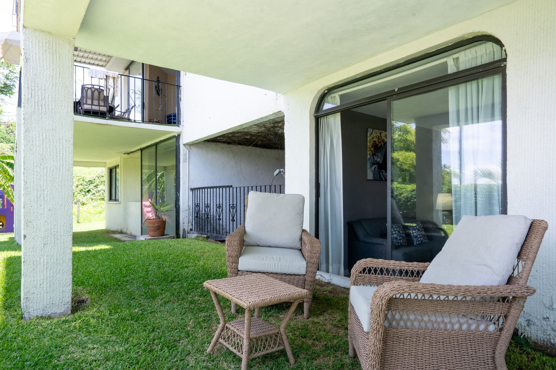 Patio con muebles de mimbre sobre césped verde, debajo de un edificio blanco con puerta corrediza de vidrio.