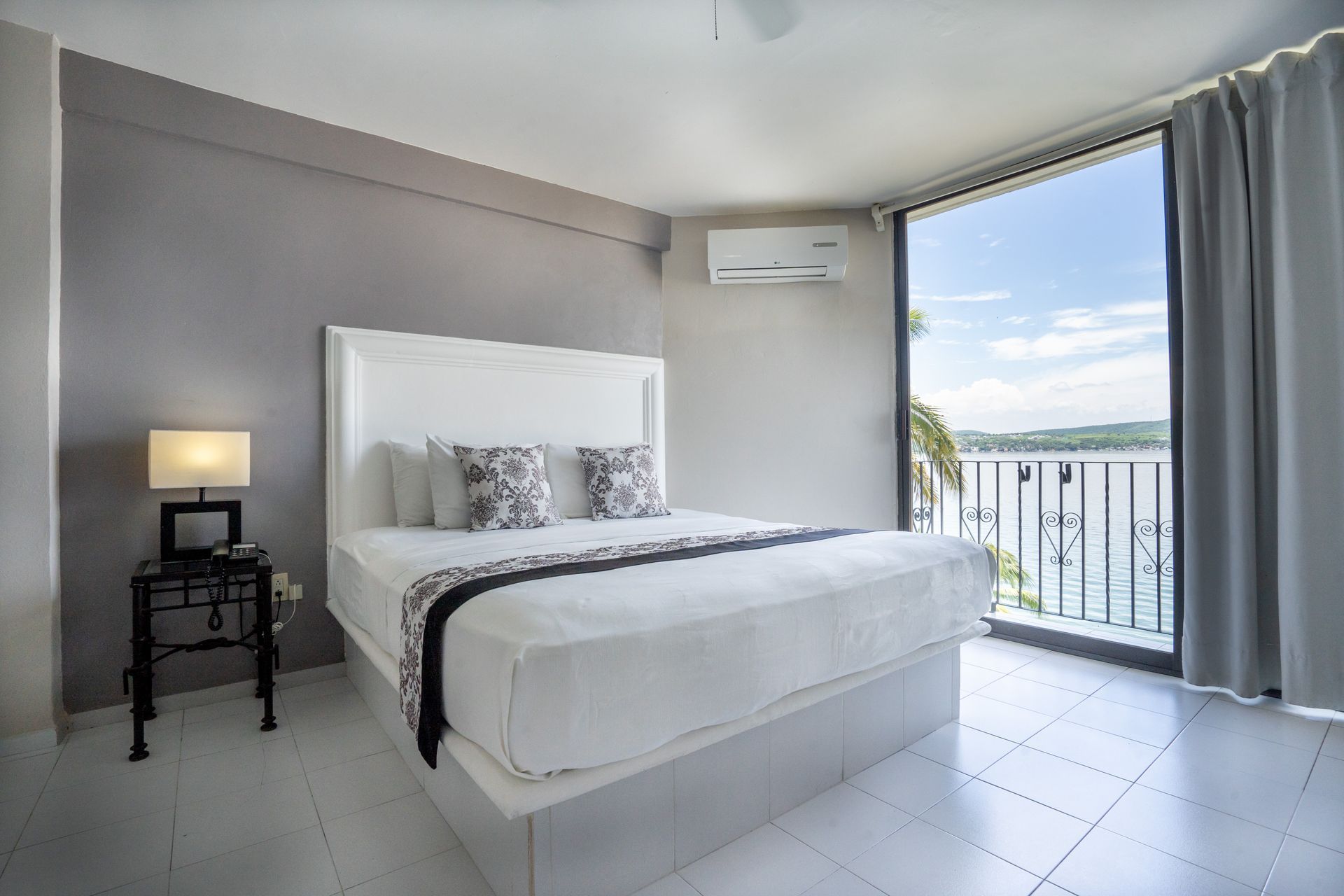 Dormitorio con cama blanca, pared gris y vista al mar desde la ventana.