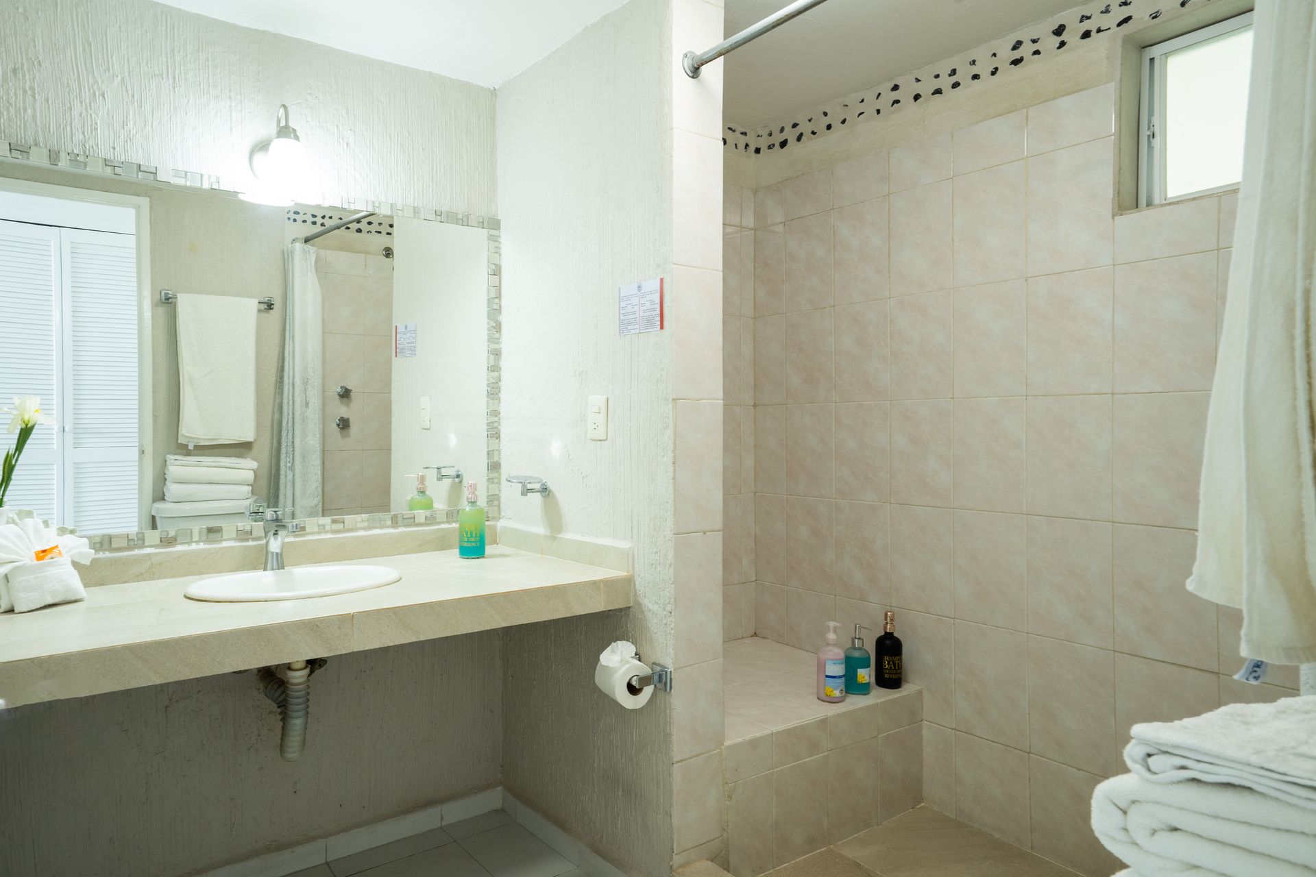 Baño con azulejos blancos y beige, lavabo accesible y ducha con pequeño banco.