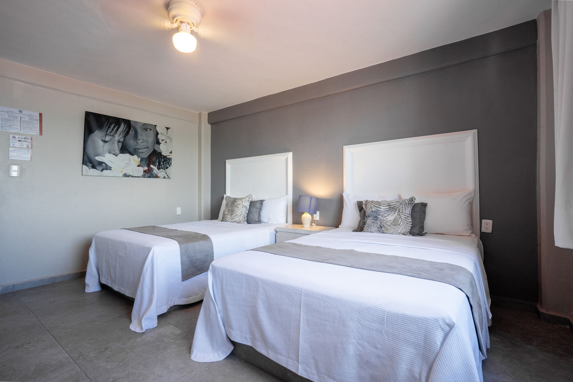 Dos camas individuales en una habitación, ropa de cama blanca con detalles grises, pared gris, obras de arte.