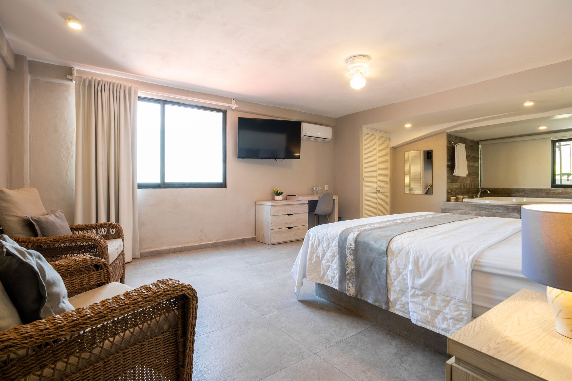 Amplia habitación de hotel con cama, zona de estar, escritorio, TV y baño privado. Colores neutros y luz natural.