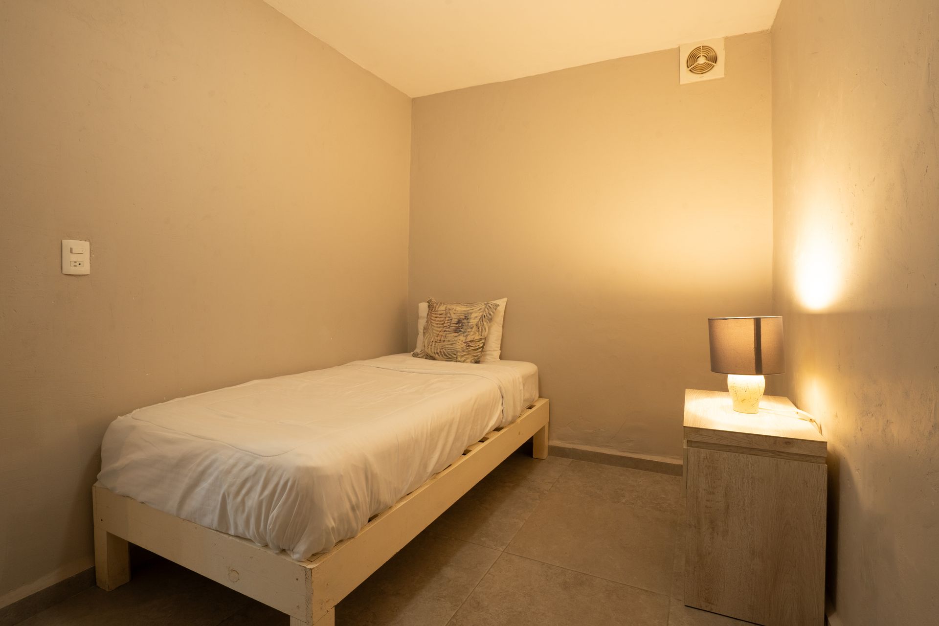 Dormitorio pequeño y minimalista con cama individual, mesita de noche y lámpara; paredes beige, luz tenue.