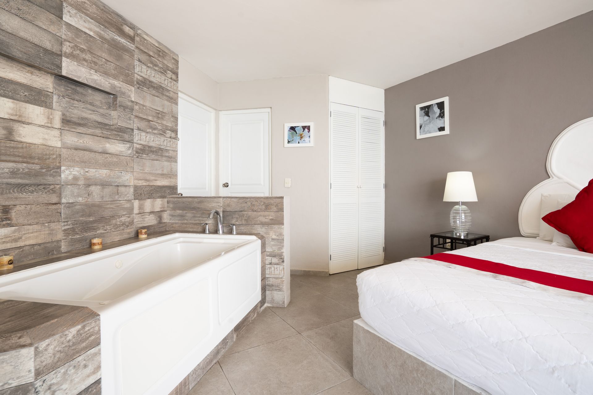 Dormitorio con bañera blanca, pared de piedra gris, cama con almohadas rojas y una mesa auxiliar con lámpara.
