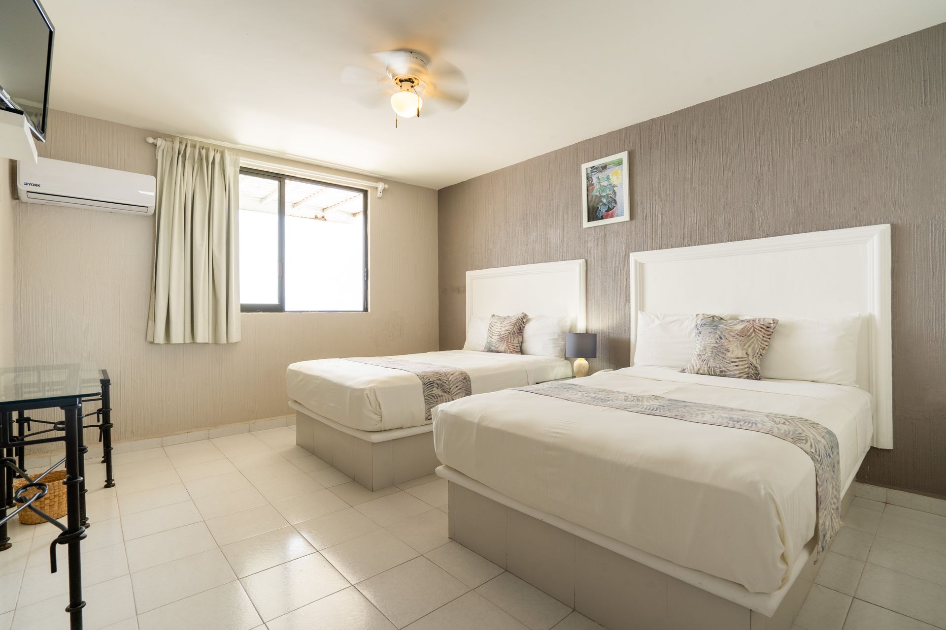 Habitación de hotel con dos camas blancas, paredes neutrales, ventana, ventilador de techo y TV.