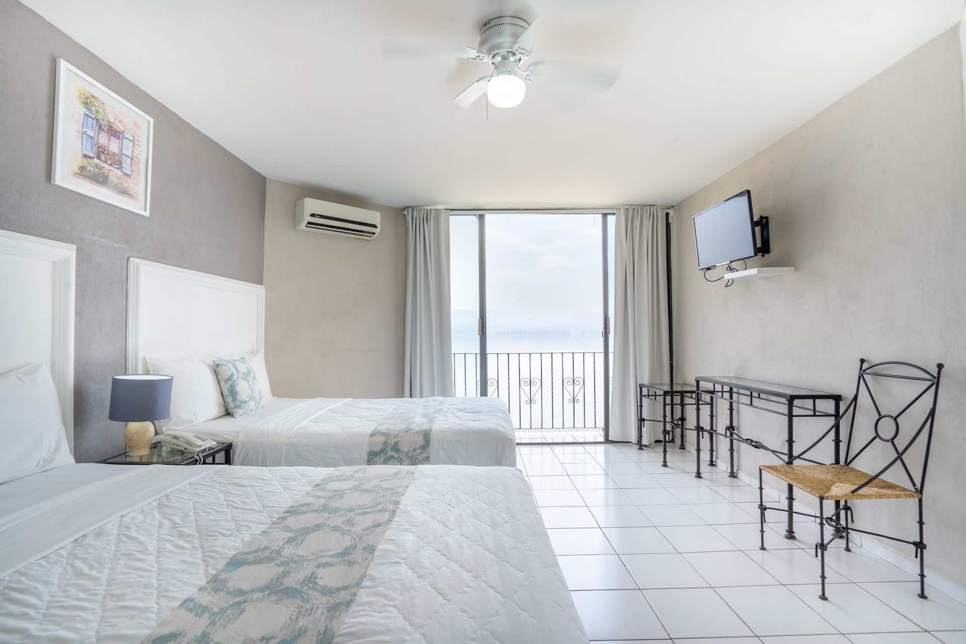 Recámara con dos camas, vista al mar, decoración en blanco y gris, TV y ventilador de techo.