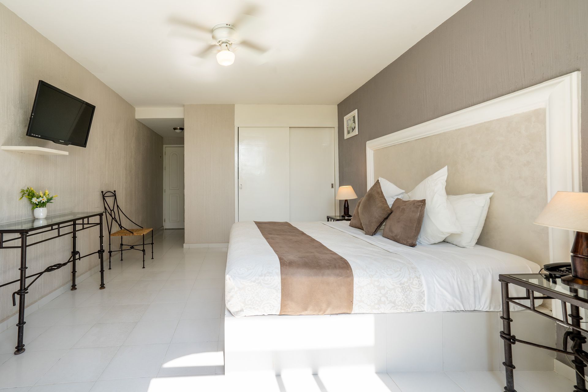 Recámara con cama King, TV, mesa decorativa, pisos blancos y paredes grises.