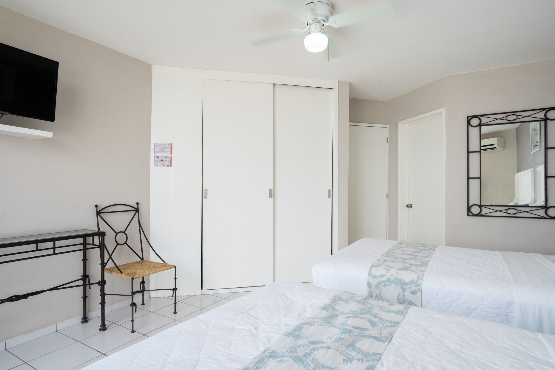 Dormitorio con dos camas, armario, silla, escritorio, televisor y espejo. Colores blanco, gris y negro.