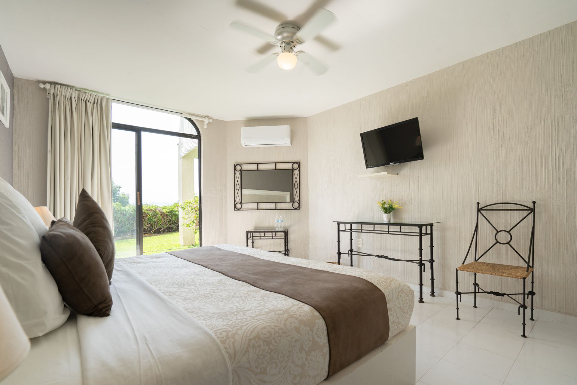 Dormitorio con cama grande, TV, sillón y una ventana que da a un césped verde.