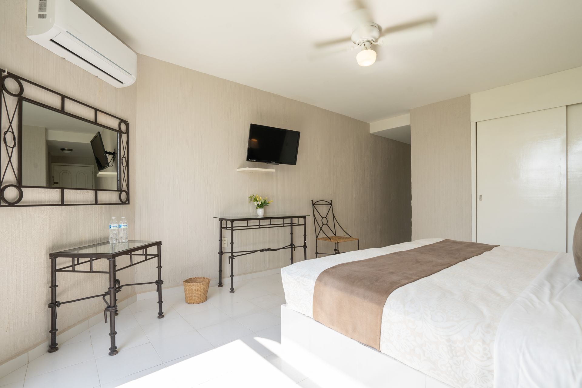 Dormitorio con cama, televisor, espejo y escritorio. Paredes y suelo blancos.